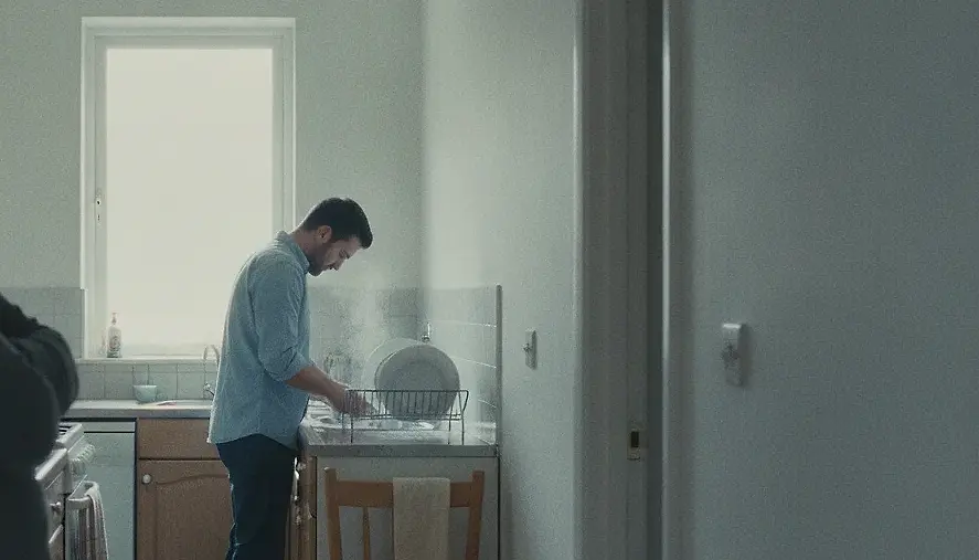 A man washing up at a kitchen sink with his head down, seen past the shoulder of someone standing in the doorway behind him.